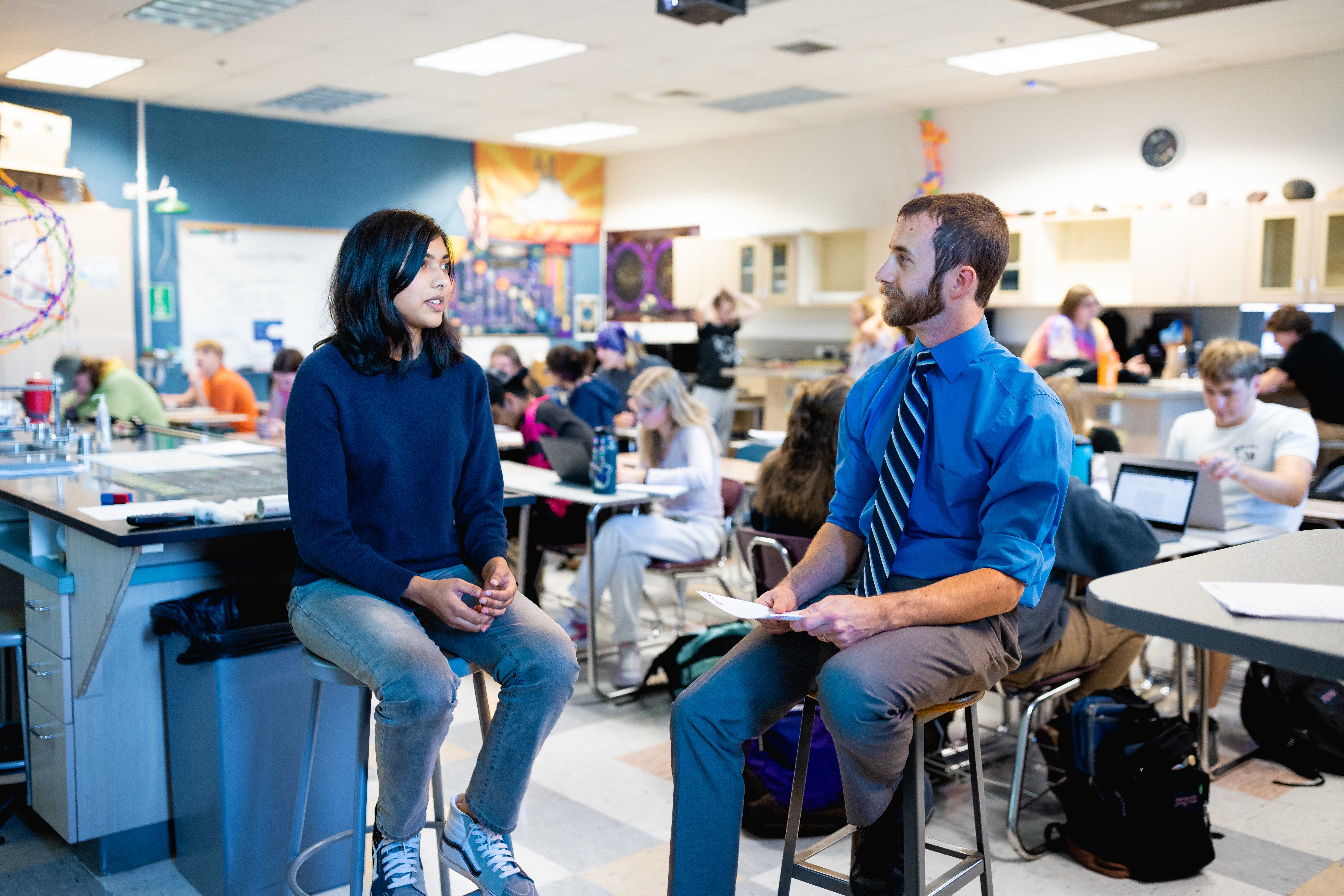 Teacher sitting with student in a classroom talking one on one with students in background